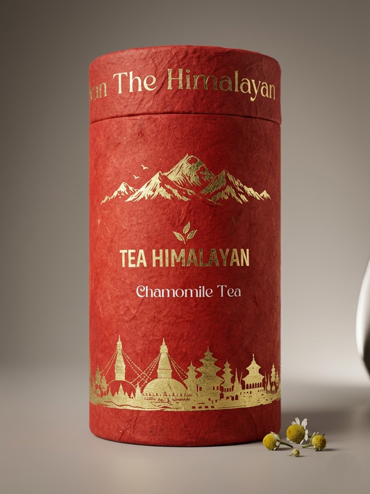 A cylindrical red tea tin labeled 'The Himalayan' and 'TEA HIMALAYAN Chamomile Tea' with gold mountain and city illustrations, shown on a neutral gray background with a few chamomile flowers beside it.